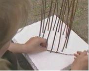 Kuvassa on kiinnitetty pajunoksia styrox-levyyn ympyrän muotoon ja tekijä aloittaa pajuamppelin punonnan alhaalta käsin kiertämällä pajunoksaa pystyssä olevien pajunoksien ympärille